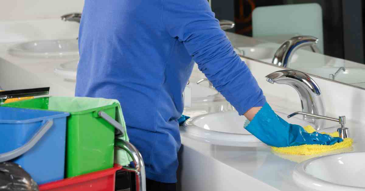 A janitor wiping down a restroom sink area with a yellow towel. A janitor’s cart with buckets is behind the person.