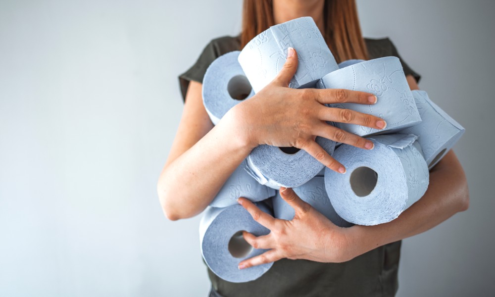 Woman holding a pile of toilet paper rolls in her arms. The toilet paper is blue and the wall behind her is gray.