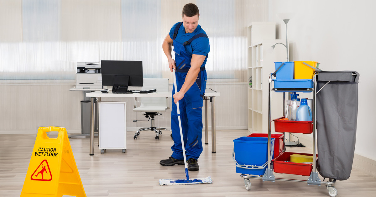 A janitor cleaning an office floor. He’s near a cart with cleaning supplies and a 